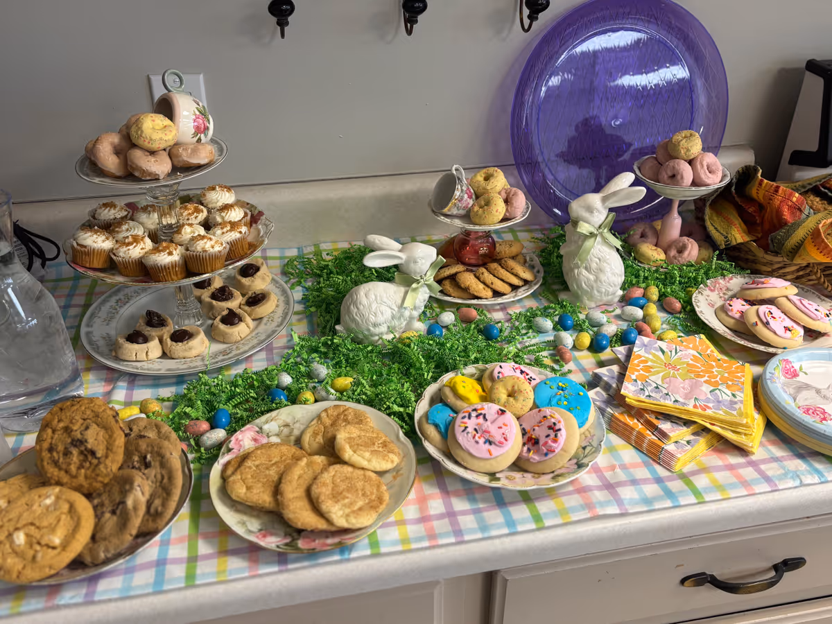 A festive dessert table with various cookies, cupcakes, and donuts displayed on plates and tiered stands. The table is decorated with green shredded paper, colorful candy eggs, and two white ceramic bunny figurines with green ribbons around their necks. There are also floral-patterned napkins and plates on the table, set on a pastel plaid tablecloth.