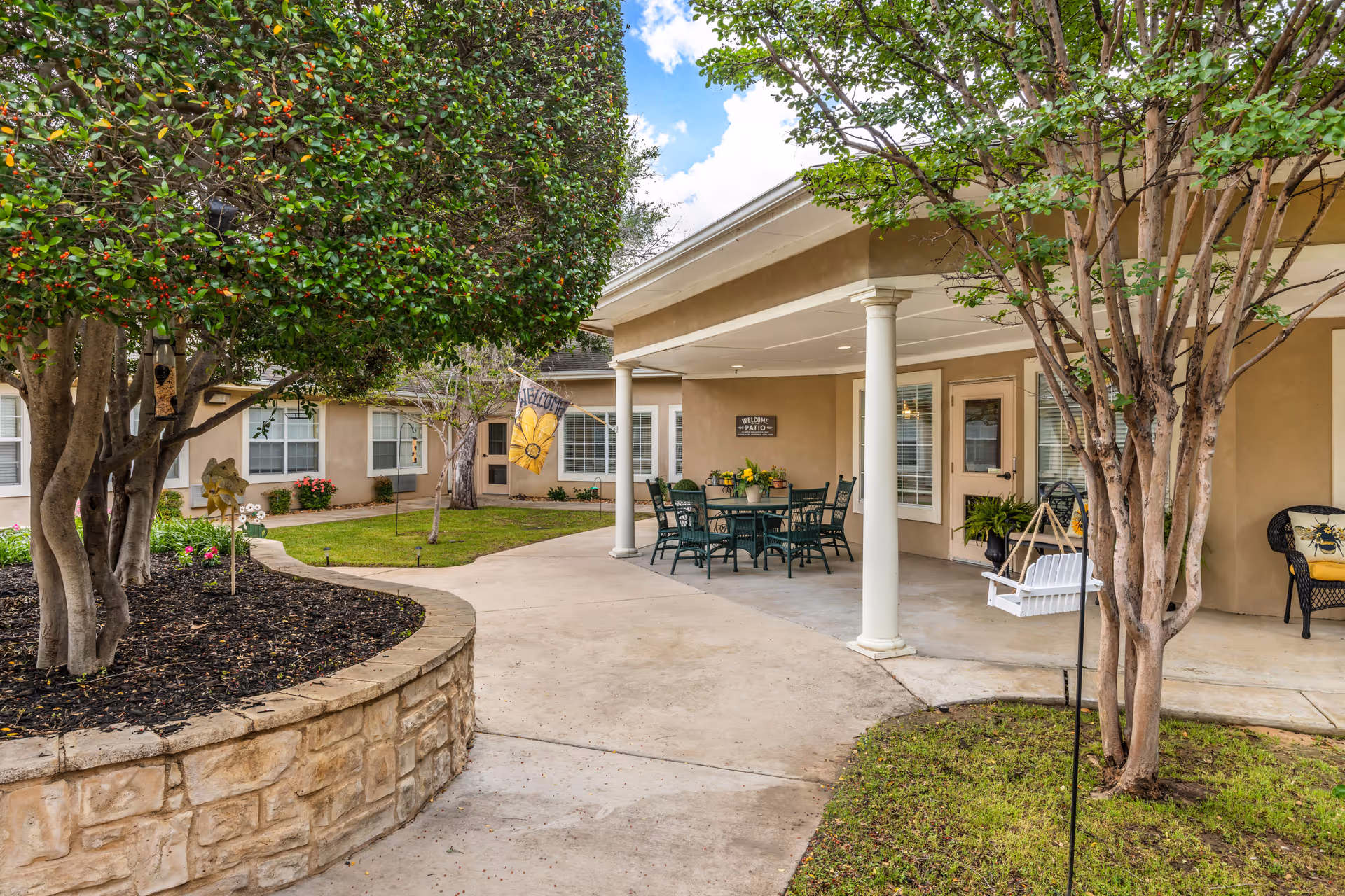 Outdoor patio area at Brookdale Oak Hollow featuring a covered seating area with a table and chairs, a white hanging swing, and surrounding greenery including trees and shrubs. A stone planter and a welcome flag are also visible.