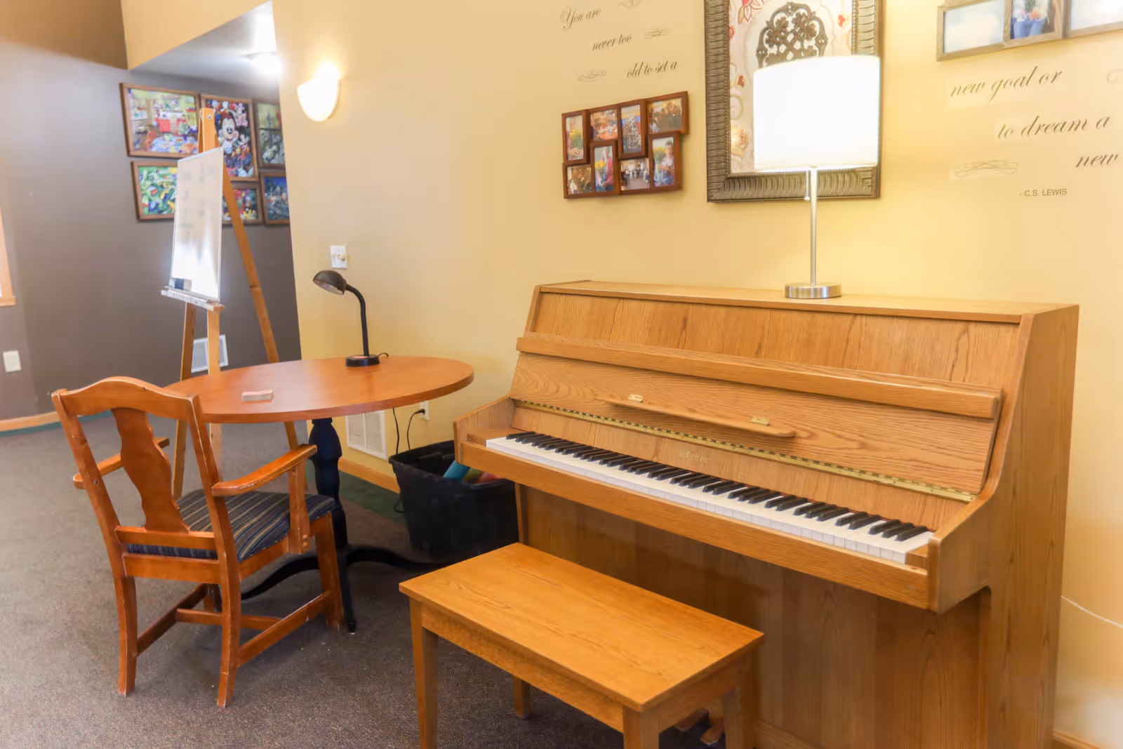 A cozy interior corner featuring a wooden upright piano with a matching bench, a small round wooden table with a chair, a desk lamp, and a wall lamp. The wall behind the piano has framed photos, a decorative mirror, and a quote by C.S. Lewis. In the background, there is a whiteboard on an easel and colorful artwork on the walls.