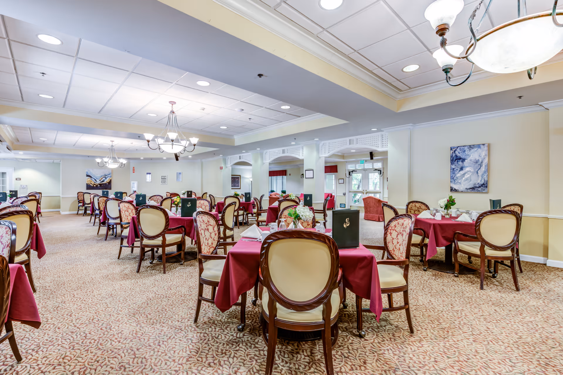 Spacious dining room in a senior living facility with multiple tables covered in burgundy tablecloths, each set with menus, napkins, and floral centerpieces. The room features patterned carpet, elegant chairs with cream and floral upholstery, soft yellow walls, and ceiling chandeliers providing warm lighting. Artwork is displayed on the walls, and large windows allow natural light to enter.