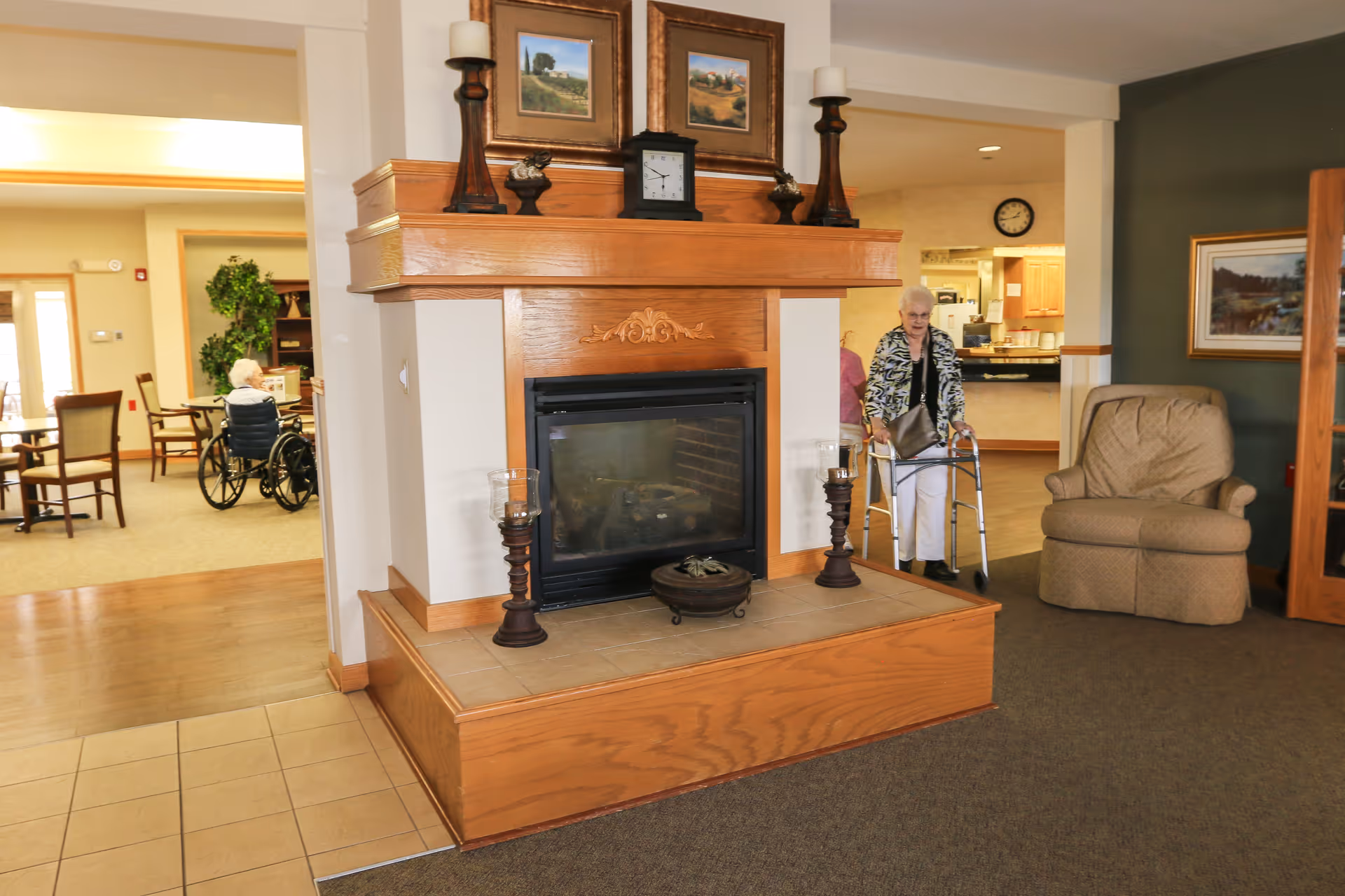 Interior view of a senior living facility showing a wooden fireplace with decorative items and framed pictures on the mantel. To the right, an elderly woman using a walker is walking near a beige armchair. In the background to the left, another elderly person in a wheelchair is seated at a table in a dining area with chairs and plants.
