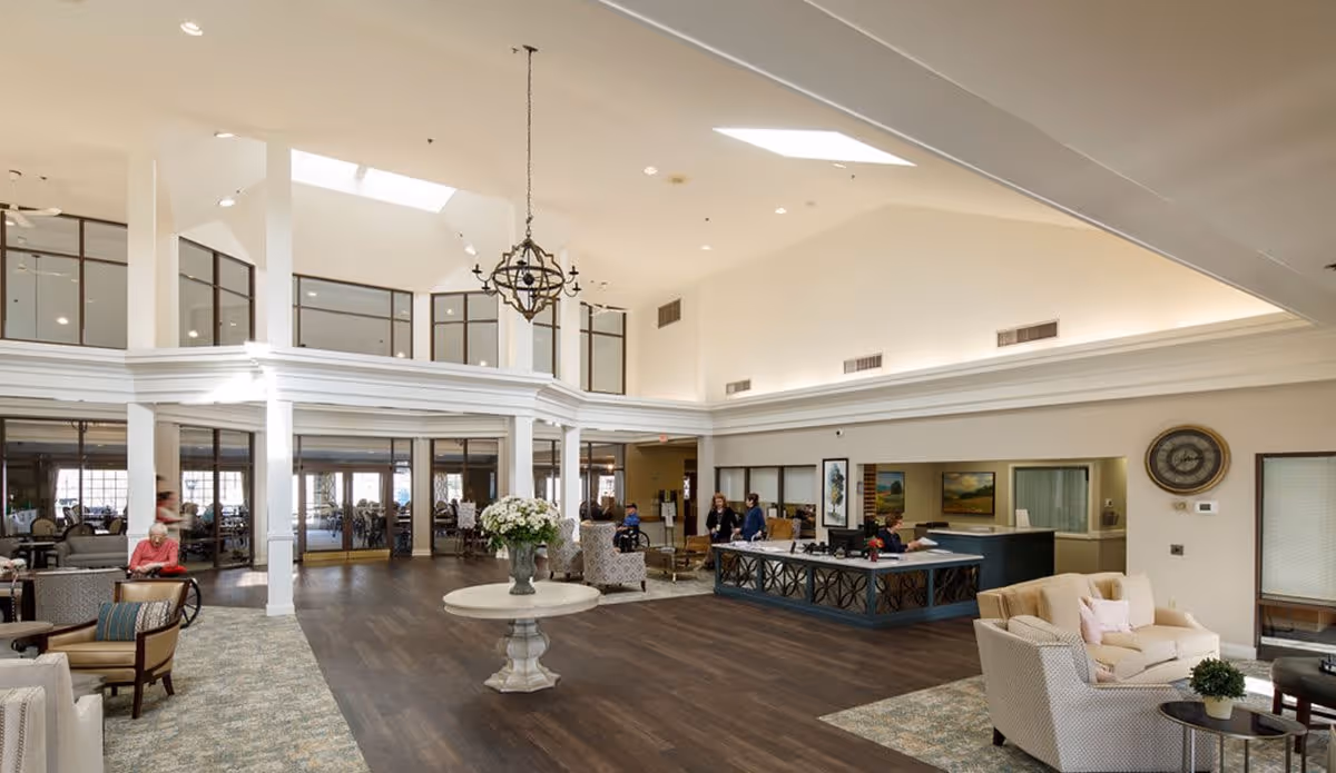 Spacious and well-lit senior living facility lobby with high ceilings, large windows, and a chandelier. The room features a central round table with a flower arrangement, a reception desk with staff, comfortable seating areas with sofas and chairs, and several seniors sitting and walking around.