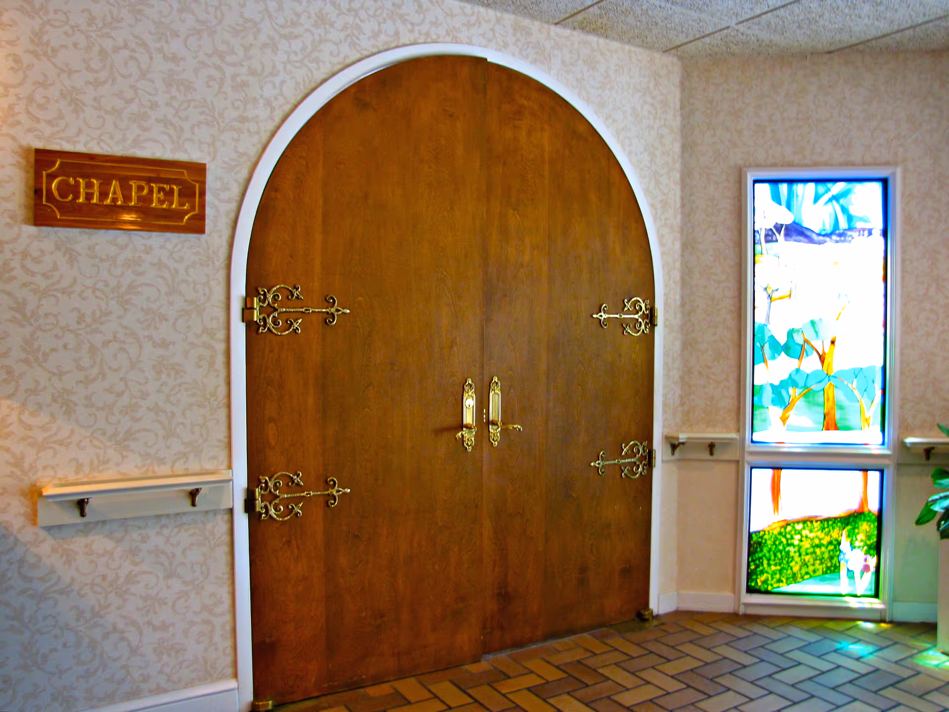 Double wooden doors with ornate gold hinges and handles set in an arched doorway. To the left of the doors is a wooden sign that reads 'CHAPEL'. To the right is a tall stained glass window depicting a colorful outdoor scene with trees and greenery. The walls have a light patterned wallpaper and the floor is tiled with a brick pattern.