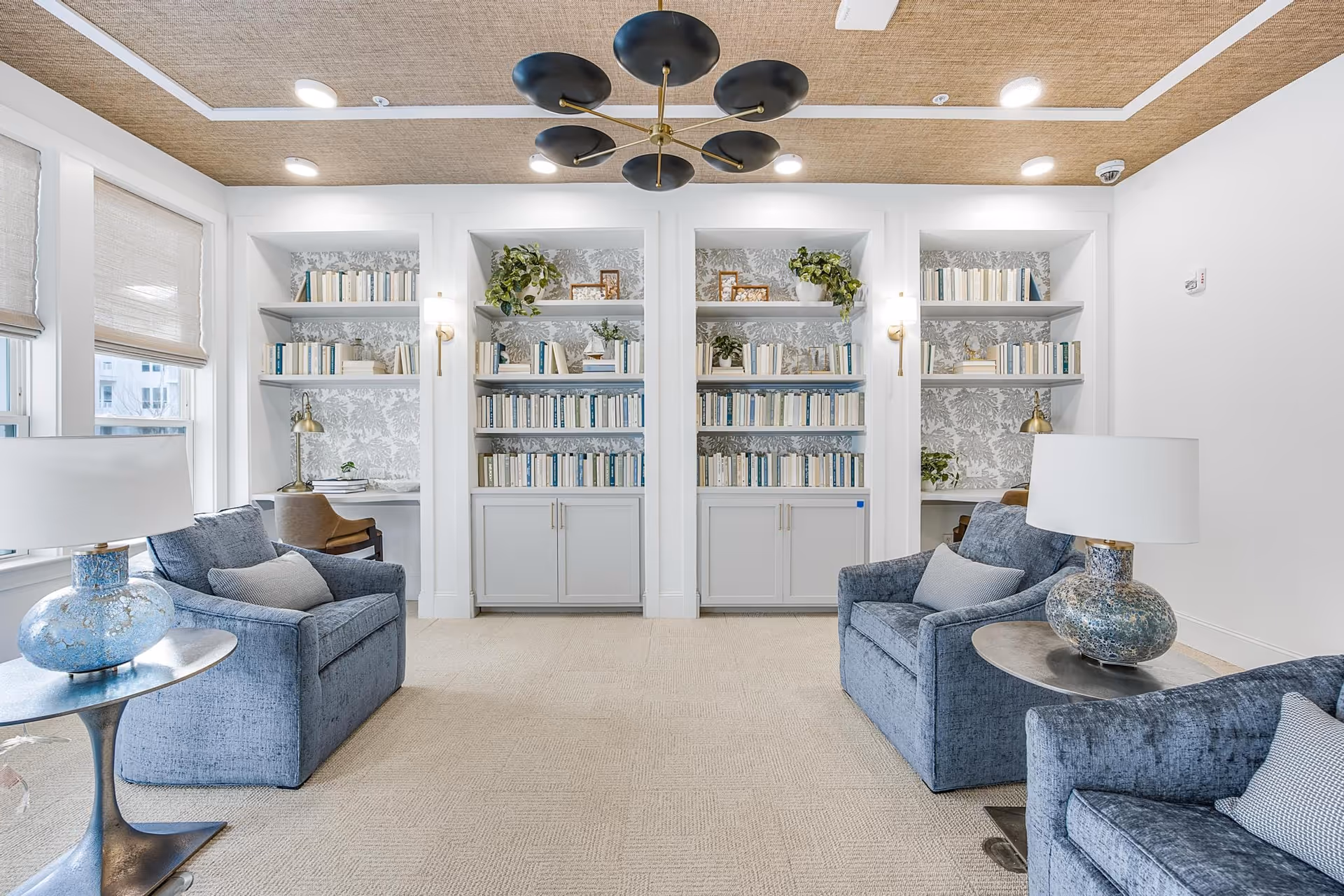 A bright and cozy living room area featuring two blue upholstered armchairs with gray cushions, two side tables with decorative lamps, built-in white bookshelves filled with books and plants, and two small desks with chairs near the windows. The ceiling has a textured design with recessed lighting and a modern black and gold chandelier.