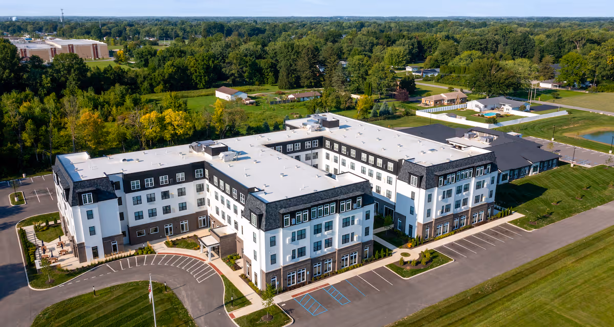Aerial view of a large, modern senior living facility named Vita of Marion, surrounded by green lawns, trees, and parking areas. The building is white with dark roofing and multiple windows, set in a spacious, suburban environment with clear skies.