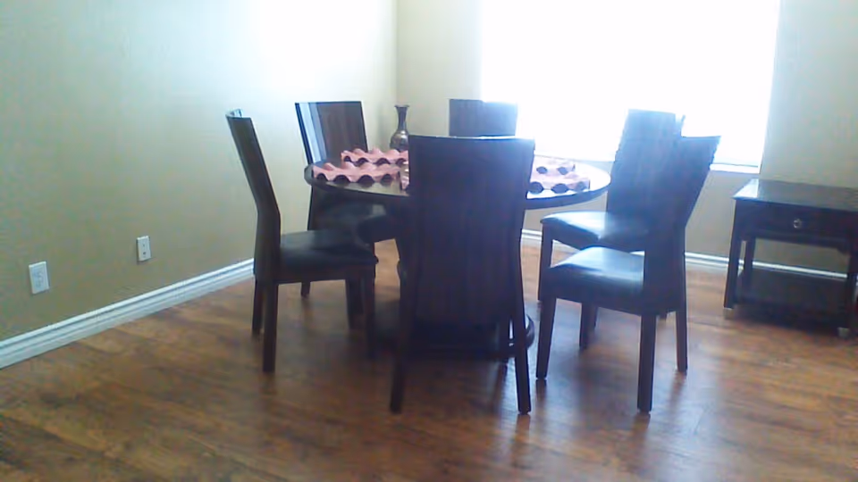 A dining area with a round wooden table surrounded by six dark wooden chairs with black cushions. The table has a decorative centerpiece and a pink table runner. The room has wooden flooring, beige walls, and a window letting in natural light. There is also a small dark wooden side table against the wall.