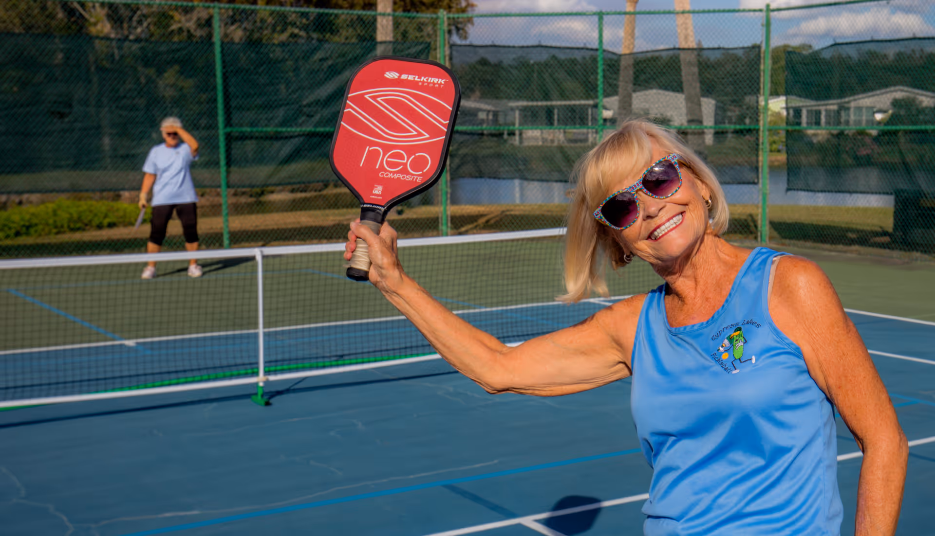 An elderly woman wearing sunglasses and a blue sleeveless shirt is smiling and holding a red pickleball paddle on an outdoor pickleball court. Another person is standing on the opposite side of the net, preparing to play. The court is surrounded by a green fence with a lake and buildings visible in the background.