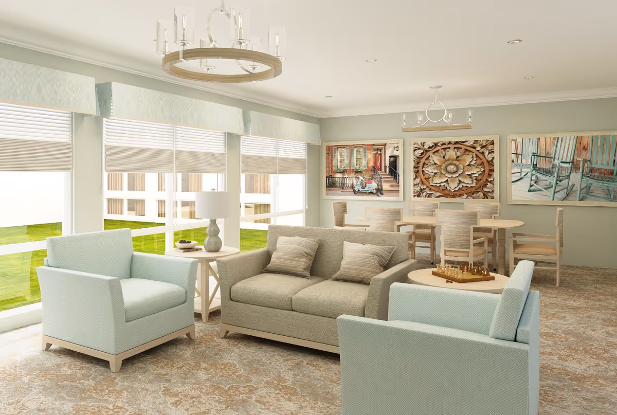 A bright and spacious living room area with large windows letting in natural light. The room features a beige loveseat with two striped pillows, two light blue armchairs, a round wooden coffee table with a chess set, and a side table with a lamp and decorative bowl. In the background, there is a dining table with six chairs and three large framed pictures on the wall depicting a scooter by a building, an ornate floral carving, and green rocking chairs on a porch. The room has light green walls, patterned carpet, and two modern chandeliers.