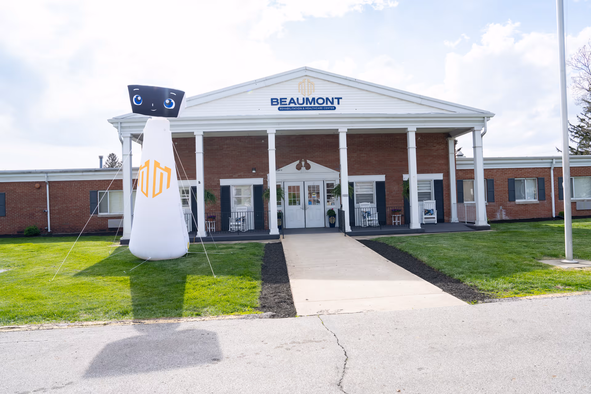 Front exterior view of Beaumont Rehabilitation and Healthcare Center, a single-story brick building with white columns and a covered entrance. There is a large inflatable figure with a smiling face and blue eyes on the lawn to the left of the entrance. The sky is partly cloudy.