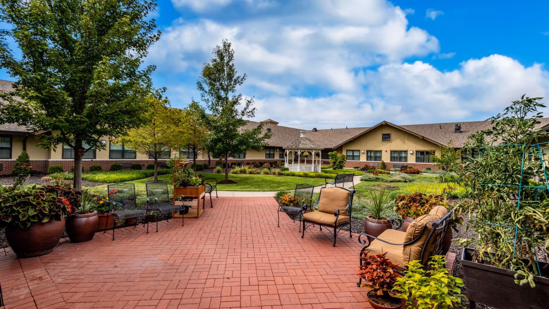 Outdoor patio area with brick flooring, several cushioned chairs, metal chairs, and large potted plants. In the background, there is a well-maintained garden with trees, shrubs, and a white gazebo. The building of the facility is visible behind the garden under a partly cloudy blue sky.