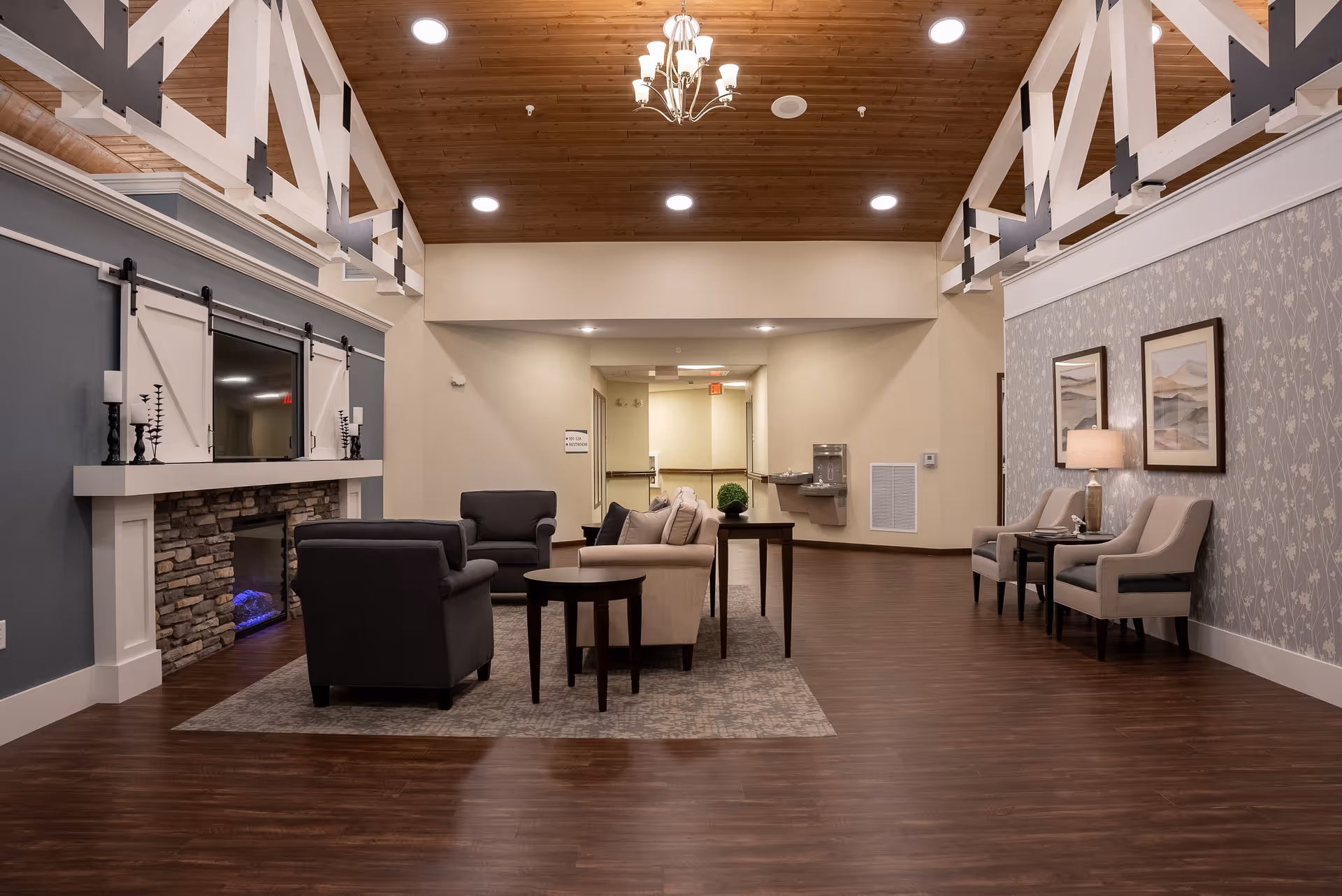 A spacious senior living facility common area with a high wooden ceiling and recessed lighting. The room features a stone fireplace with a mounted TV above it, surrounded by two black armchairs and a beige sofa arranged around a coffee table on a rug. On the right side, there are two beige armchairs with a small table and lamp between them, and two framed pictures on the wall. The floor is dark wood, and there is a water fountain and hallway visible in the background.