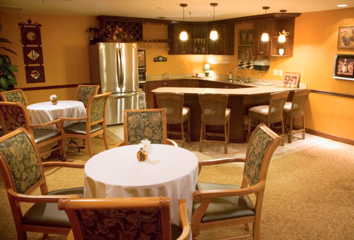 A cozy dining area with round tables covered in white tablecloths, each adorned with a small flower vase. Surrounding the tables are wooden chairs with green floral upholstery. In the background, there is a kitchen area with dark wooden cabinets, a stainless steel refrigerator, a countertop with bar stools, and warm lighting from hanging pendant lamps.