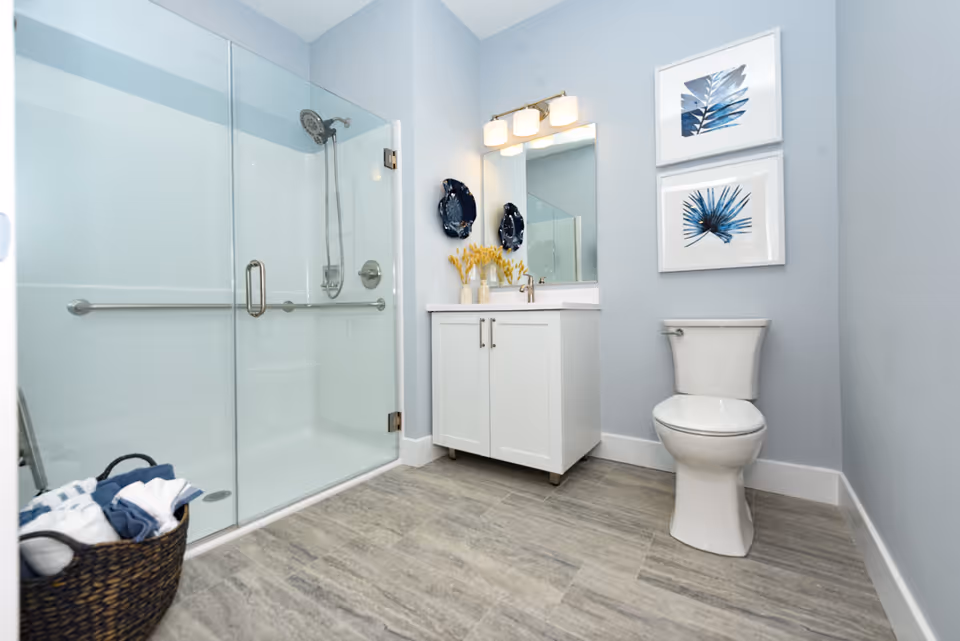 A modern bathroom with a glass-enclosed shower on the left, a white vanity with a mirror and light fixture above it in the center, and a white toilet on the right. The walls are painted light blue and there are two framed blue leaf artworks hanging above the toilet. A basket with towels is placed on the floor near the shower.