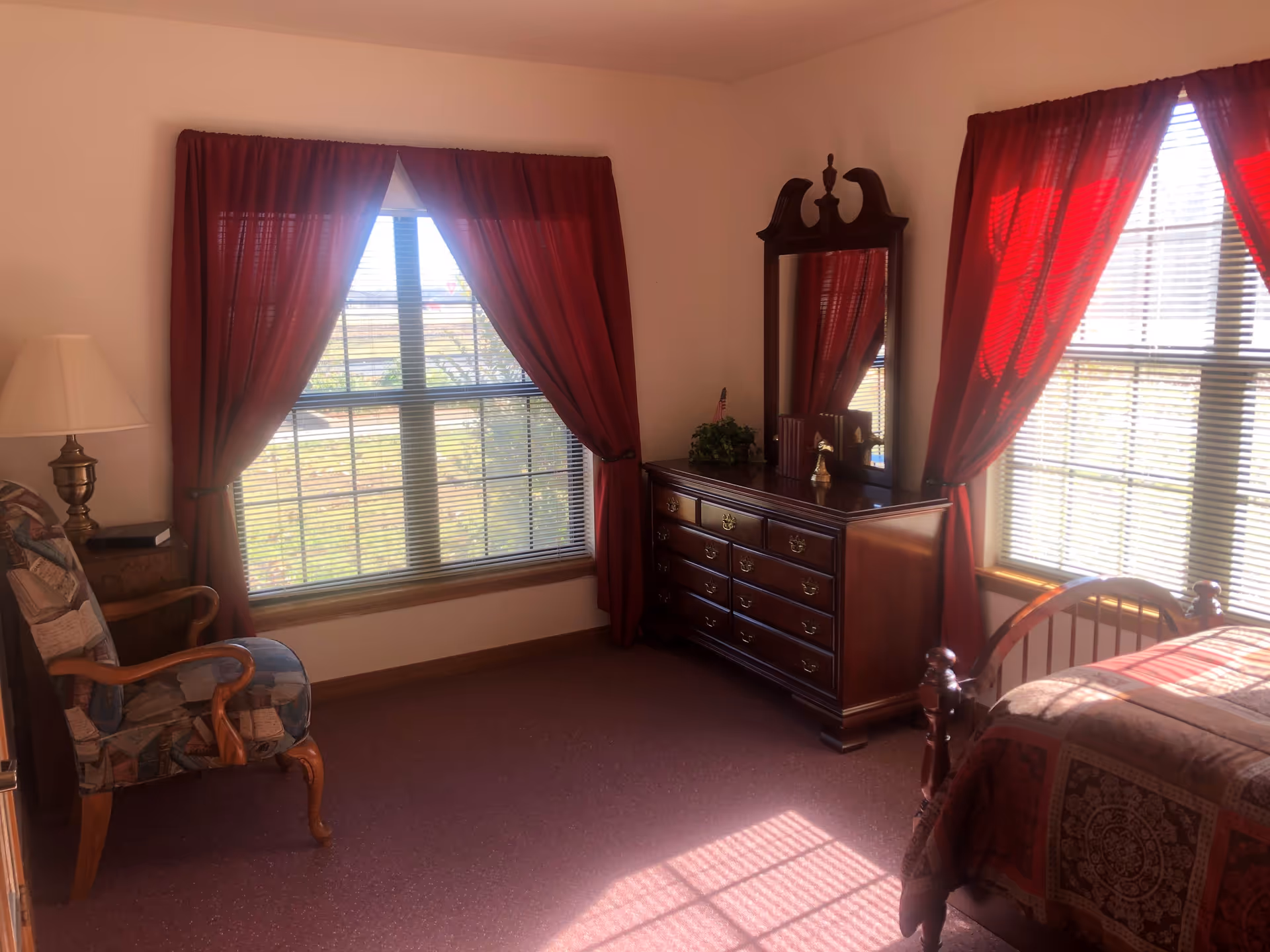 A cozy bedroom with two large windows covered by red curtains, a wooden dresser with a mirror, a patterned armchair, and a bed with a patterned quilt. Sunlight streams through the windows, casting shadows on the carpeted floor.