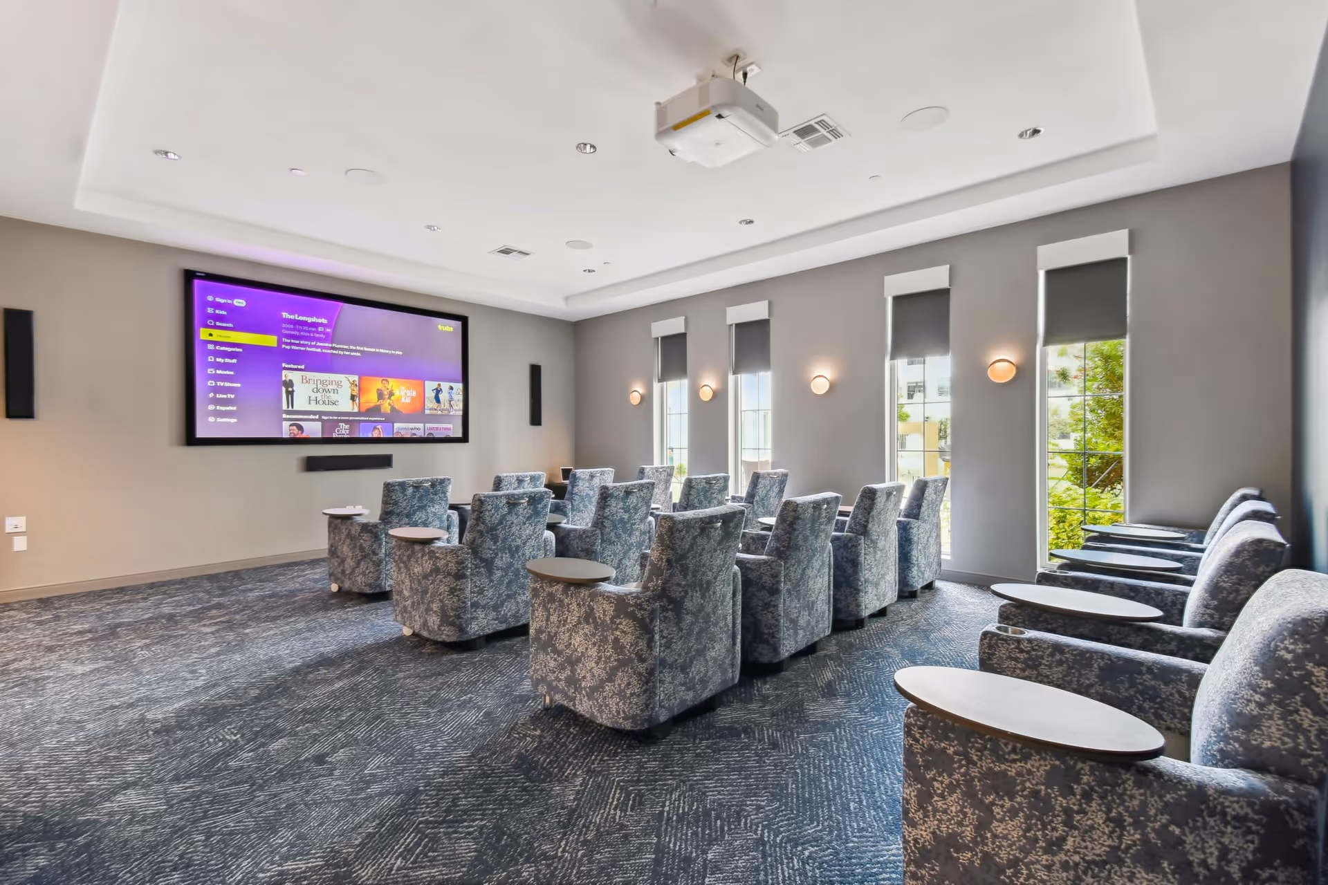 A modern media room with multiple patterned armchairs arranged in rows facing a large wall-mounted flat screen TV. The room has gray walls, three tall windows with roller shades, wall sconces, and a ceiling-mounted projector.