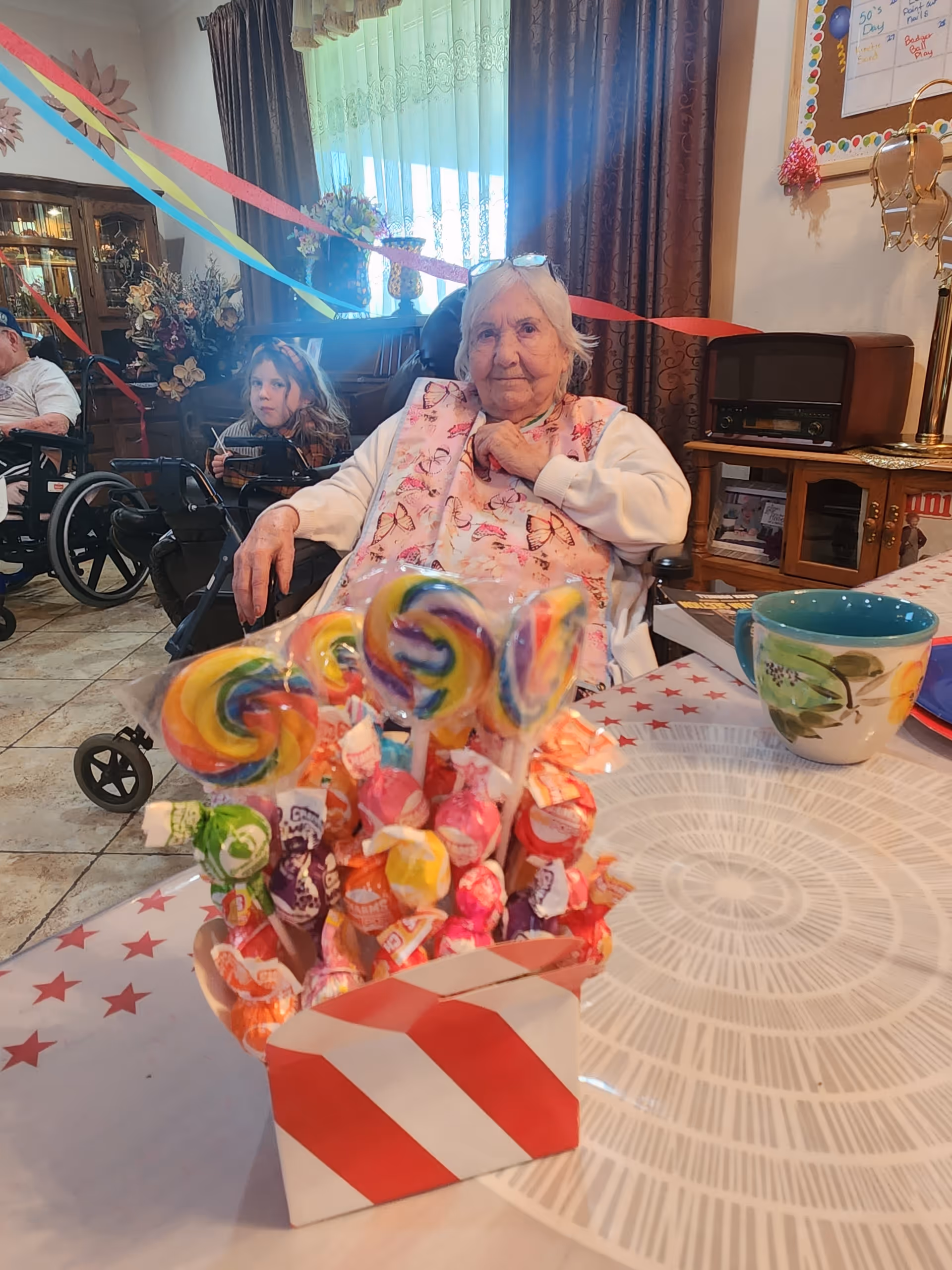 An elderly woman sitting in a wheelchair at a table decorated with a candy bouquet featuring colorful lollipops and Dum Dums. Behind her, a young girl and another person in a wheelchair are visible in a room decorated with colorful streamers and flowers. The room has tiled floors, curtains, and various furniture including a cabinet and a vintage radio.