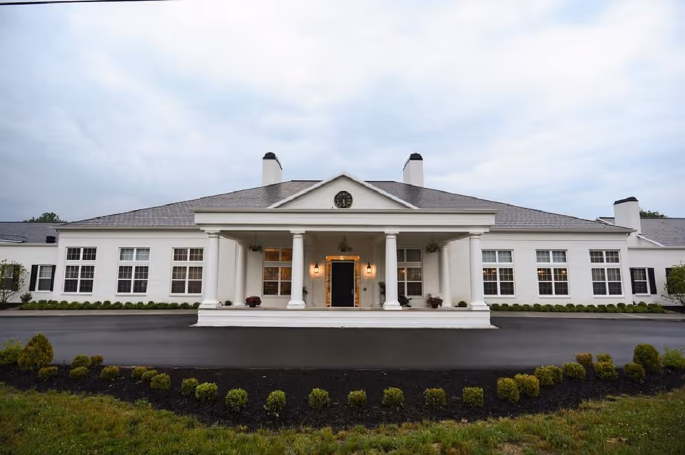 Front exterior view of Middleton Senior Living facility showing a large white building with a covered entrance supported by columns, multiple windows, and a clock above the entrance. The driveway and landscaped bushes are visible in the foreground under a cloudy sky.