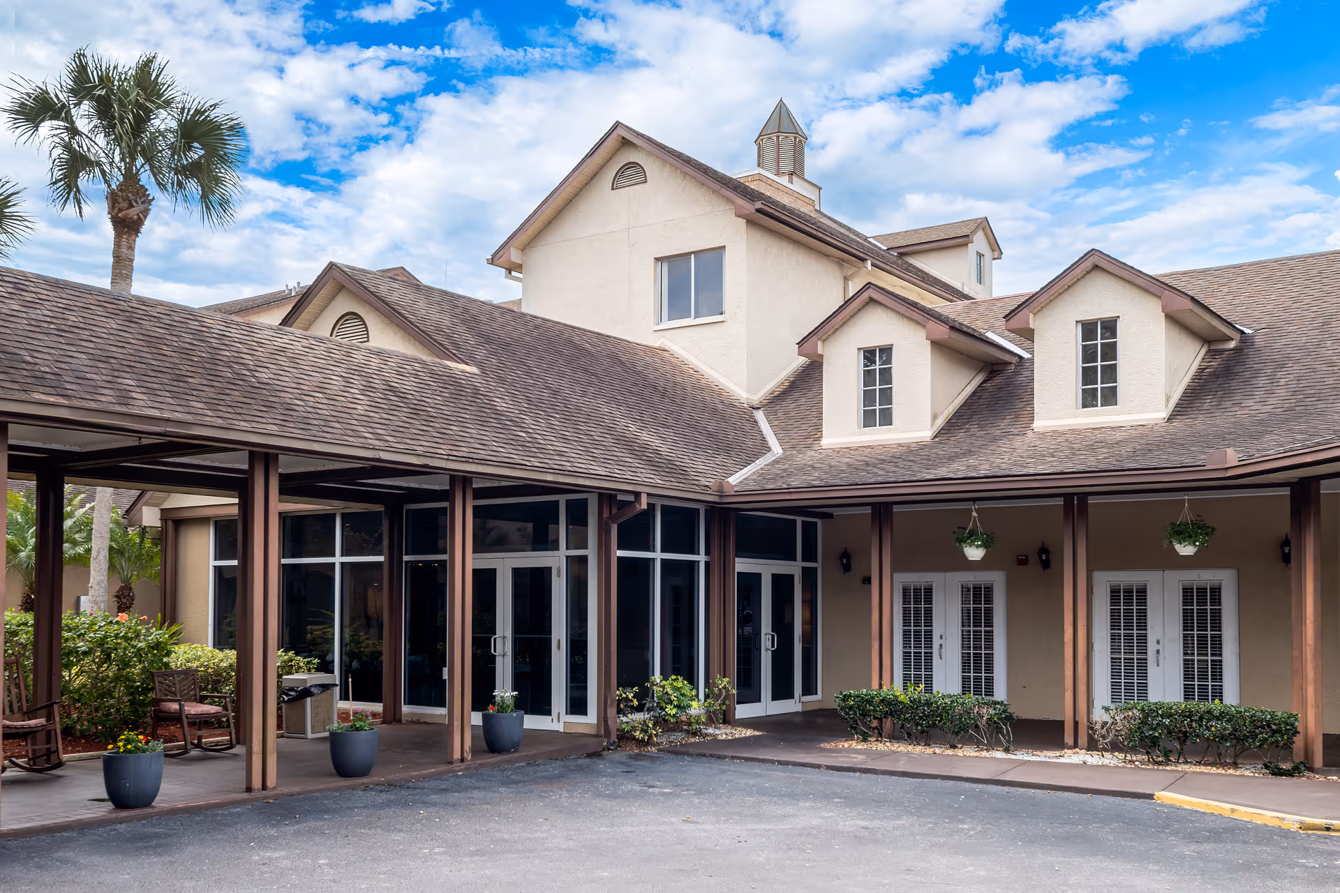 Exterior view of a senior living facility building with beige walls and brown shingled roof under a partly cloudy blue sky. The entrance area has large glass doors and windows, with a covered walkway supported by wooden pillars. There are potted plants and hanging planters near the entrance, along with some outdoor seating including rocking chairs. A tall palm tree is visible on the left side.
