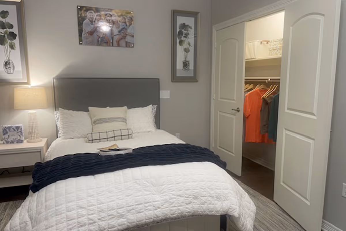 A neatly made bed with white bedding and multiple pillows in a bedroom. There is a bedside table with a lamp and a small framed picture. The walls are decorated with framed artwork and a family photo above the bed. To the right, there is an open double door leading to a walk-in closet with clothes hanging inside.