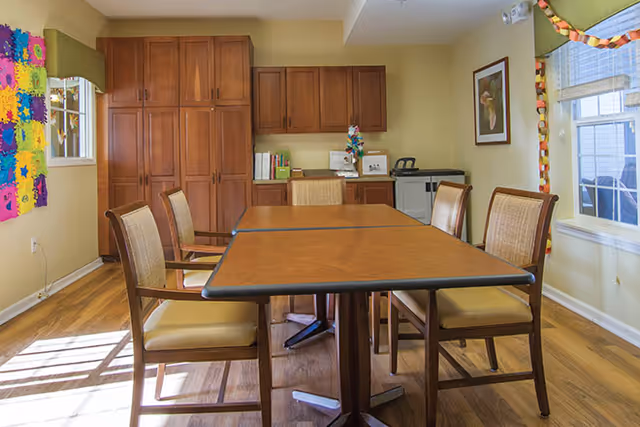 Sunlit dining area with a wooden table and chairs, built-in cabinets, and windows with colorful decorations.