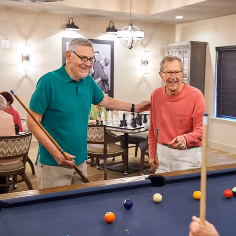 Two elderly men smiling and playing pool in a well-lit recreational room with a chess set on a table in the background and other seniors seated nearby.