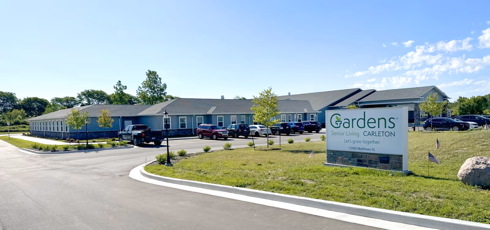 Exterior view of Gardens of Carleton senior living facility on a sunny day, showing a single-story building with multiple windows, a parking lot with several cars, a grassy area with small trees and American flags, and a large sign displaying the facility's name and address.