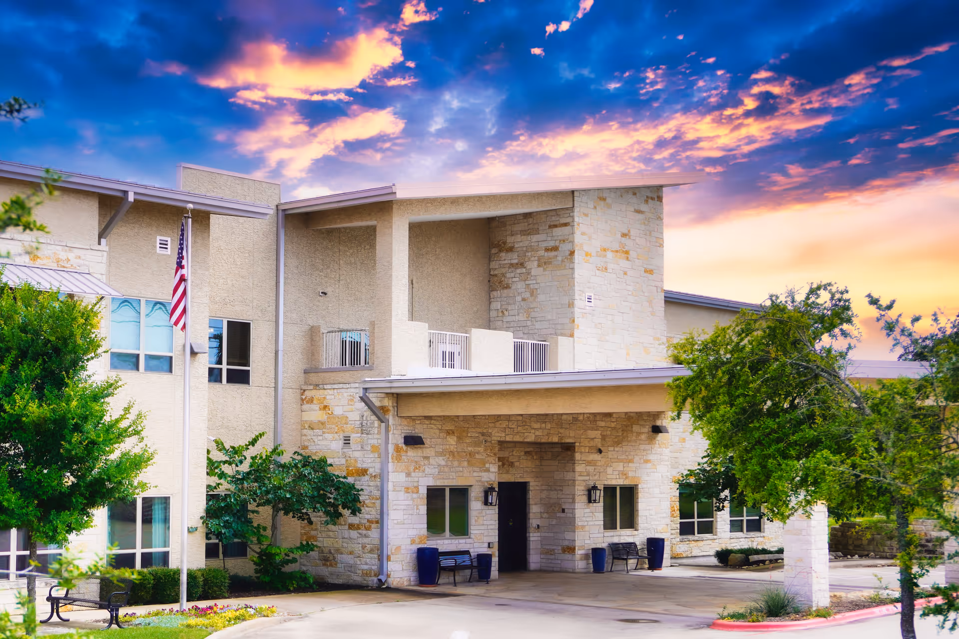 Exterior view of a senior living facility building with beige stone and stucco walls, a covered entrance, benches, and an American flag on a flagpole. The sky is colorful with a dramatic sunset.