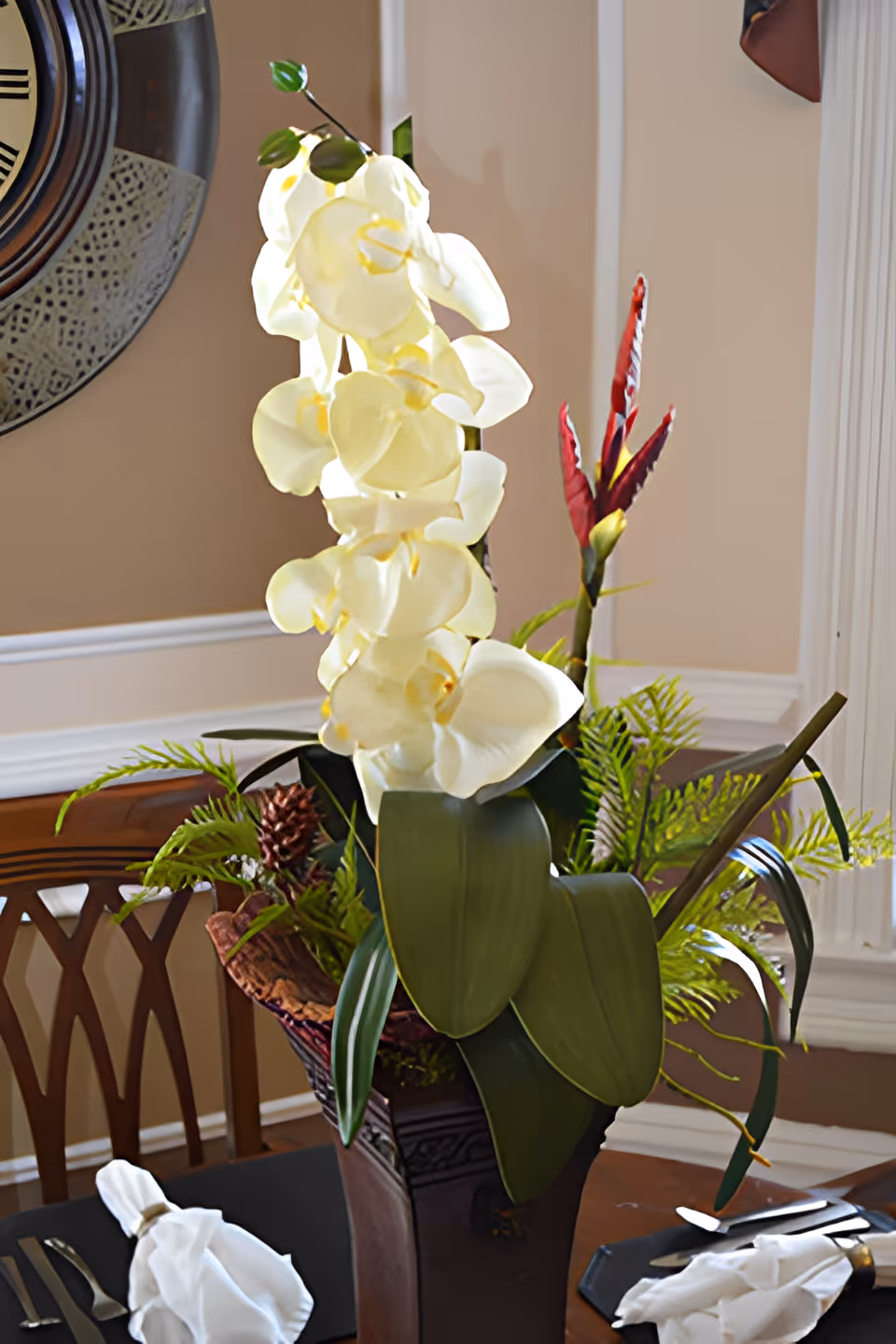 A floral centerpiece with white orchids and greenery sits on a set dining table in a dining room.