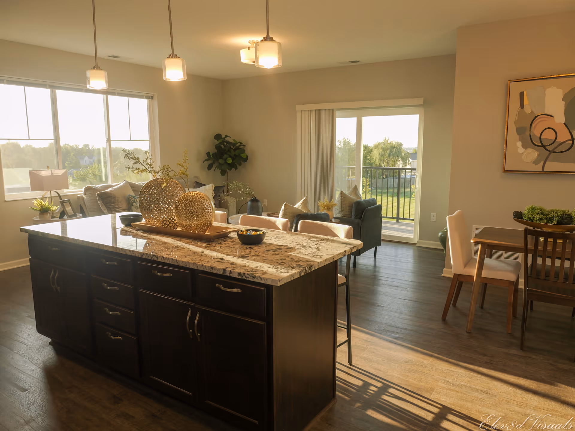 Open-concept kitchen with a large center island and bar stools opening into a sunlit living and dining area with a sliding glass door to a balcony.