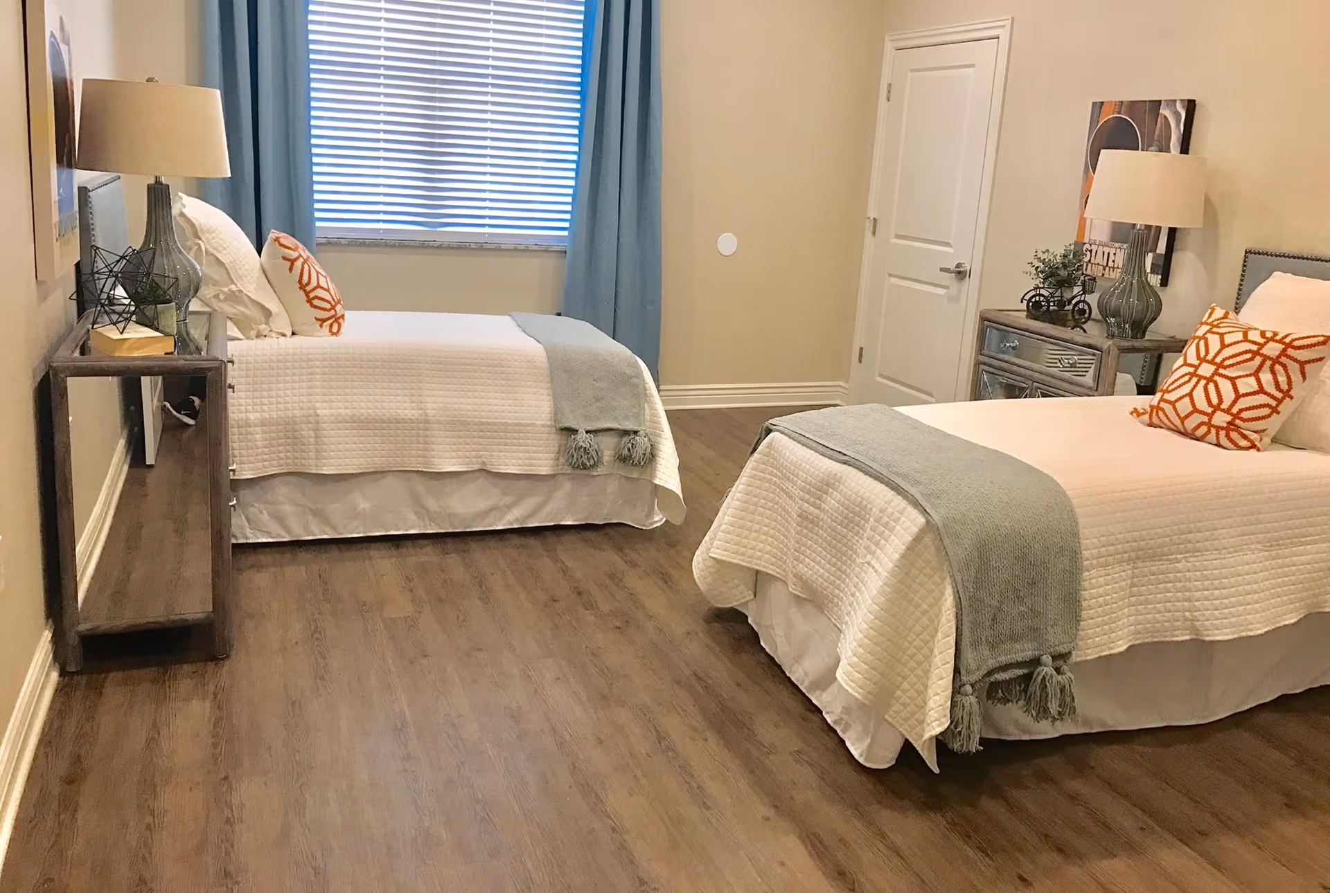 A bedroom with two twin beds, each with white quilted bedding, a light blue throw blanket, and an orange patterned pillow. There are two bedside tables with lamps and decorative items. A window with blue curtains and blinds is visible on the back wall, and a closed white door is on the right side.