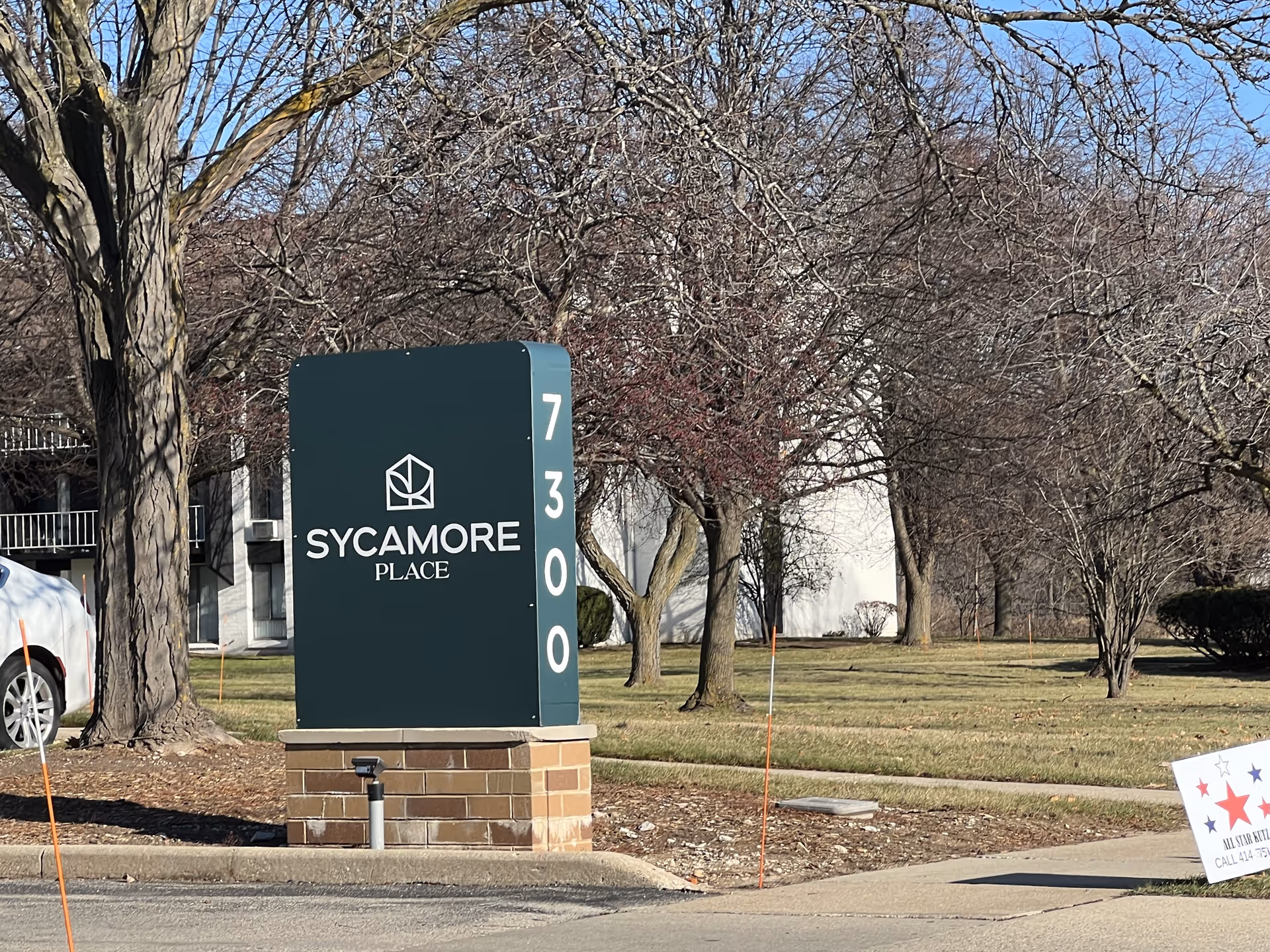 Outdoor view of a green sign with white text that reads 'Sycamore Place' and the number '7300' vertically on the side. The sign is mounted on a brick base and is located near a sidewalk with leafless trees and grass in the background. A white vehicle is partially visible on the left side.