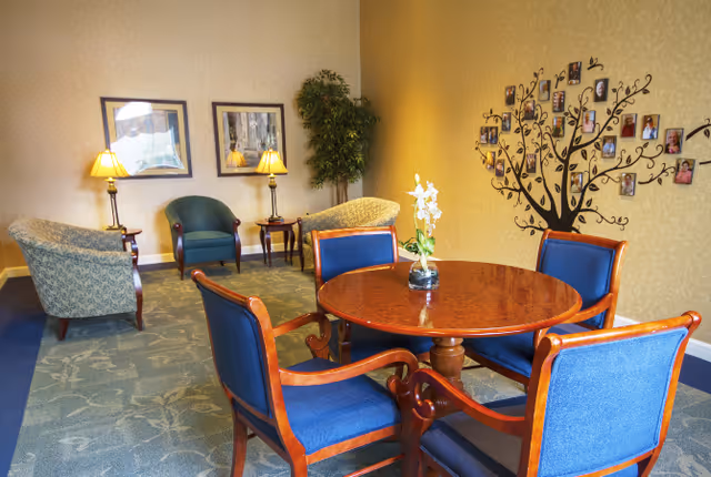 Cozy assisted-living common room with a round wooden table and four blue chairs, additional lounge chairs, lamps, a potted plant, and a photo-tree wall decoration.