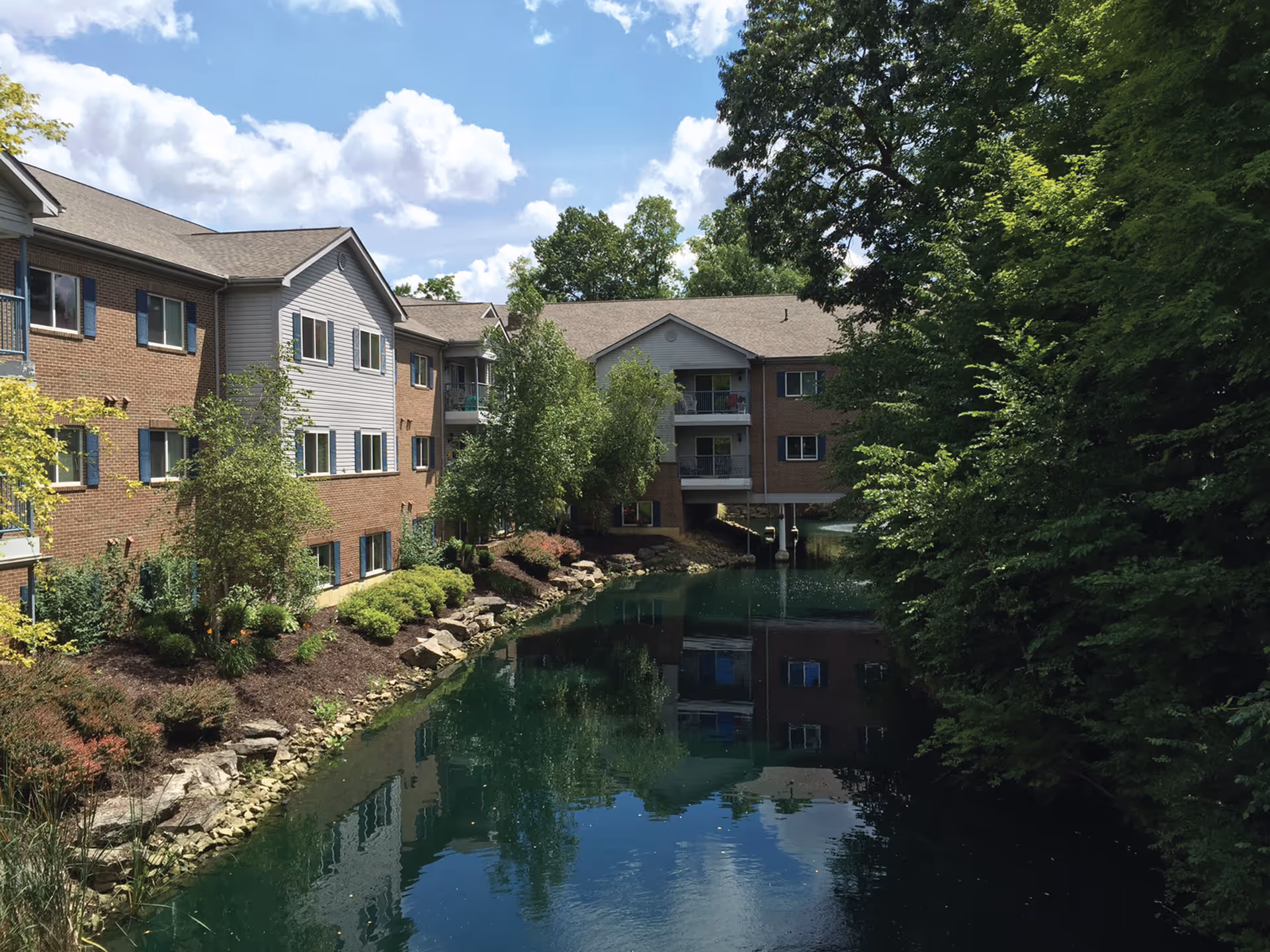 A serene outdoor view of Willow Brook Christian Village showing a brick and siding multi-story building alongside a calm pond with clear reflections of the building and surrounding trees under a partly cloudy sky.