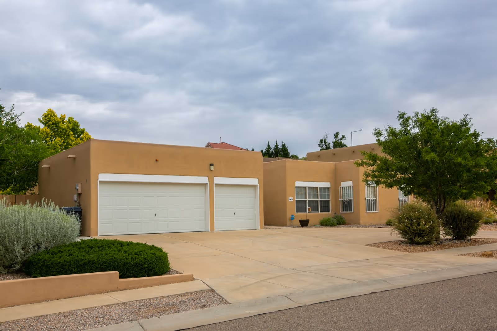 Exterior view of a single-story building with a tan stucco finish, featuring two white garage doors, several windows, and surrounded by landscaped bushes and trees under a cloudy sky.