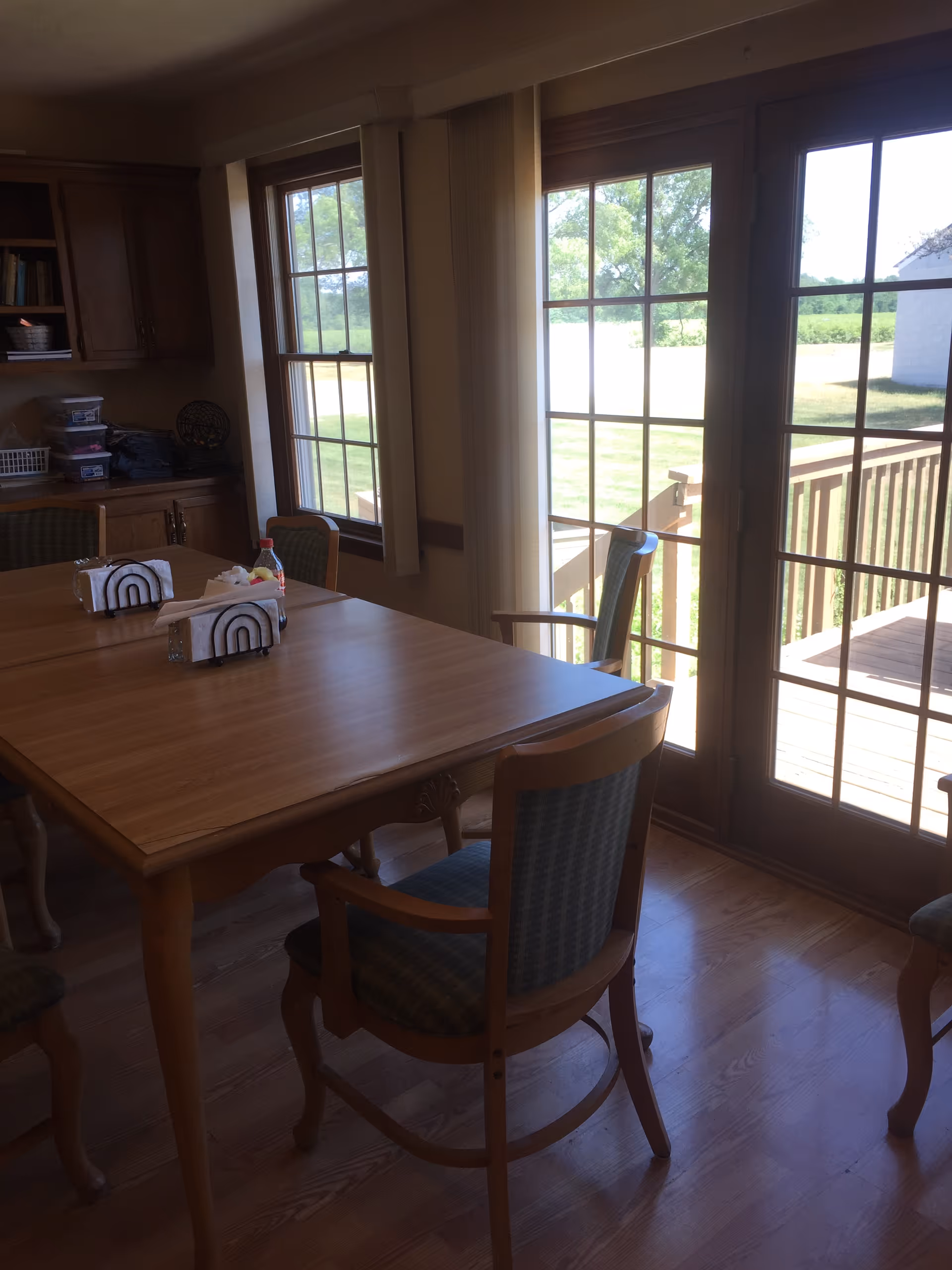 A dining area with a wooden table and several chairs with patterned upholstery. The room has large windows and glass doors that open to an outdoor deck with a view of green grass and trees. There are napkin holders and a condiment bottle on the table, and built-in wooden cabinets along one wall.