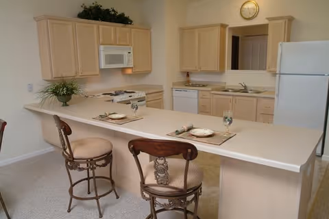 A kitchen area with light wood cabinets, white appliances including a refrigerator, microwave, stove, and dishwasher. There is a long countertop with two decorative chairs and place settings for two, including plates, napkins, and glasses. A small plant is on the counter, and a clock is mounted on the wall above the sink area.