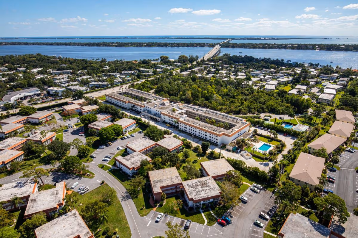 Aerial view of The Pointe at Ocean Boulevard senior living facility showing multiple residential buildings, parking lots, swimming pools, and surrounding greenery with a large body of water and a bridge in the background.