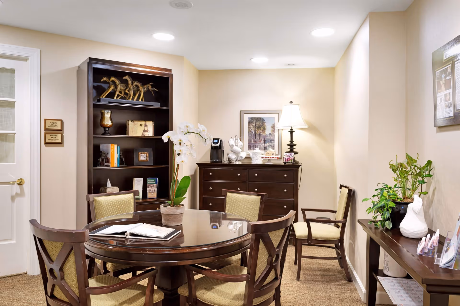A cozy interior common room with a round wooden table and chairs, bookshelf, dresser with a lamp, and potted plants.