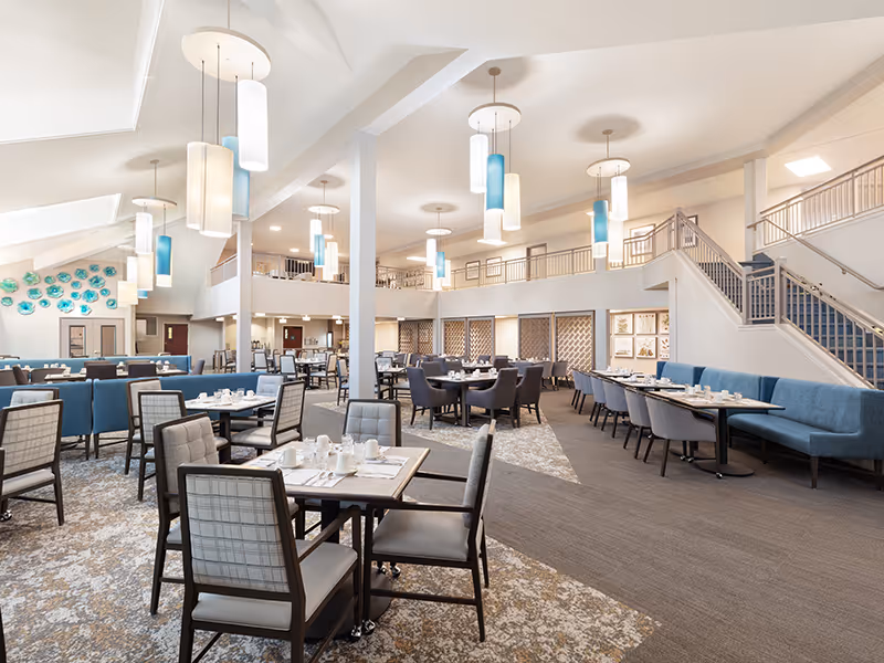 Spacious dining room in a senior living facility with multiple tables and chairs arranged neatly. The room features modern pendant lighting, a high ceiling with skylights, and a staircase leading to an upper level. The decor includes blue and gray upholstered seating and a wall adorned with decorative blue plates.