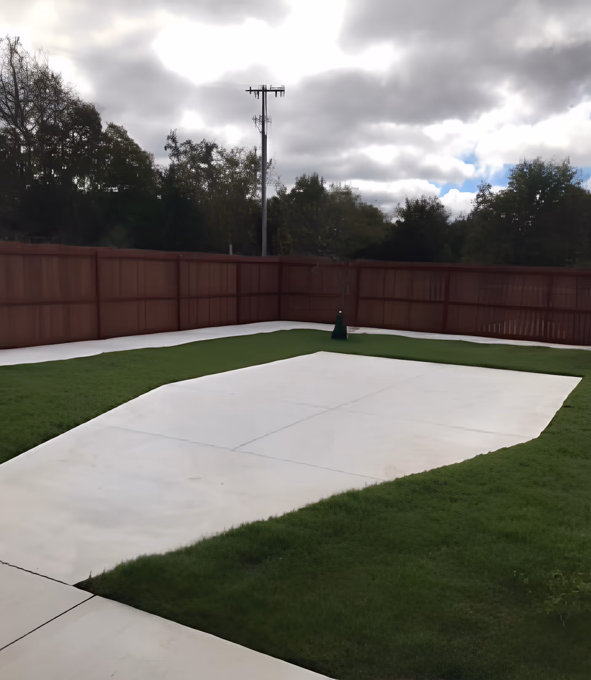 Outdoor area with a concrete pathway bordered by green grass, enclosed by a tall wooden fence. Trees and a utility pole are visible in the background under a cloudy sky.