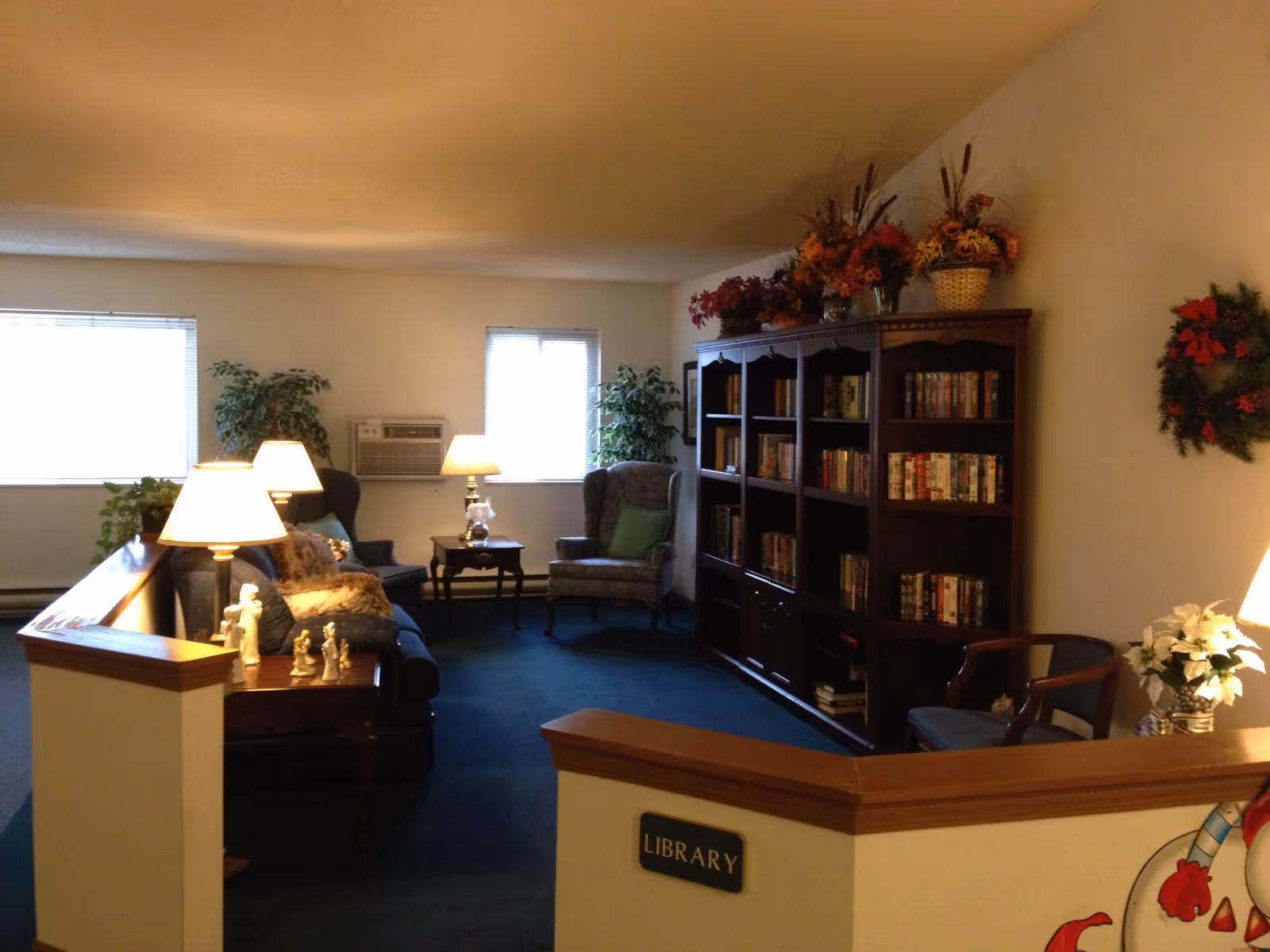 A cozy library room with a dark wooden bookshelf filled with books and decorated with flower arrangements on top. There are two armchairs, a sofa with cushions, several table lamps, and small decorative figurines on a side table. The room has two windows with blinds, green plants, and a wreath hanging on the wall. A sign on a half wall reads 'Library'.