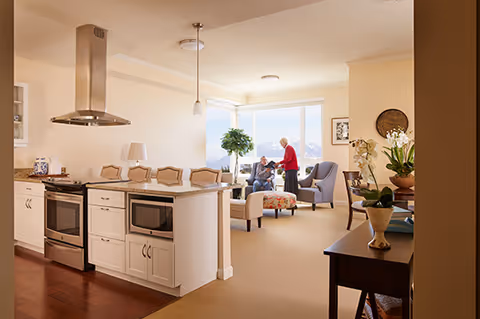 A bright and spacious interior of a senior living facility showing a modern kitchen area with white cabinets and stainless steel appliances on the left. In the background, two elderly individuals are interacting near large windows with a scenic view. The room features comfortable seating including an armchair and ottoman, with plants and decorative items adding warmth to the space.