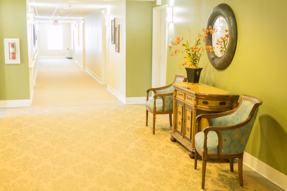 A well-lit hallway in a senior living facility with light green walls and beige patterned carpet. On the right side, there is a wooden cabinet with a vase of orange flowers on top, flanked by two upholstered chairs with blue patterned fabric. A round decorative mirror hangs above the cabinet. The hallway extends into the distance with framed pictures on the walls and a window at the far end.