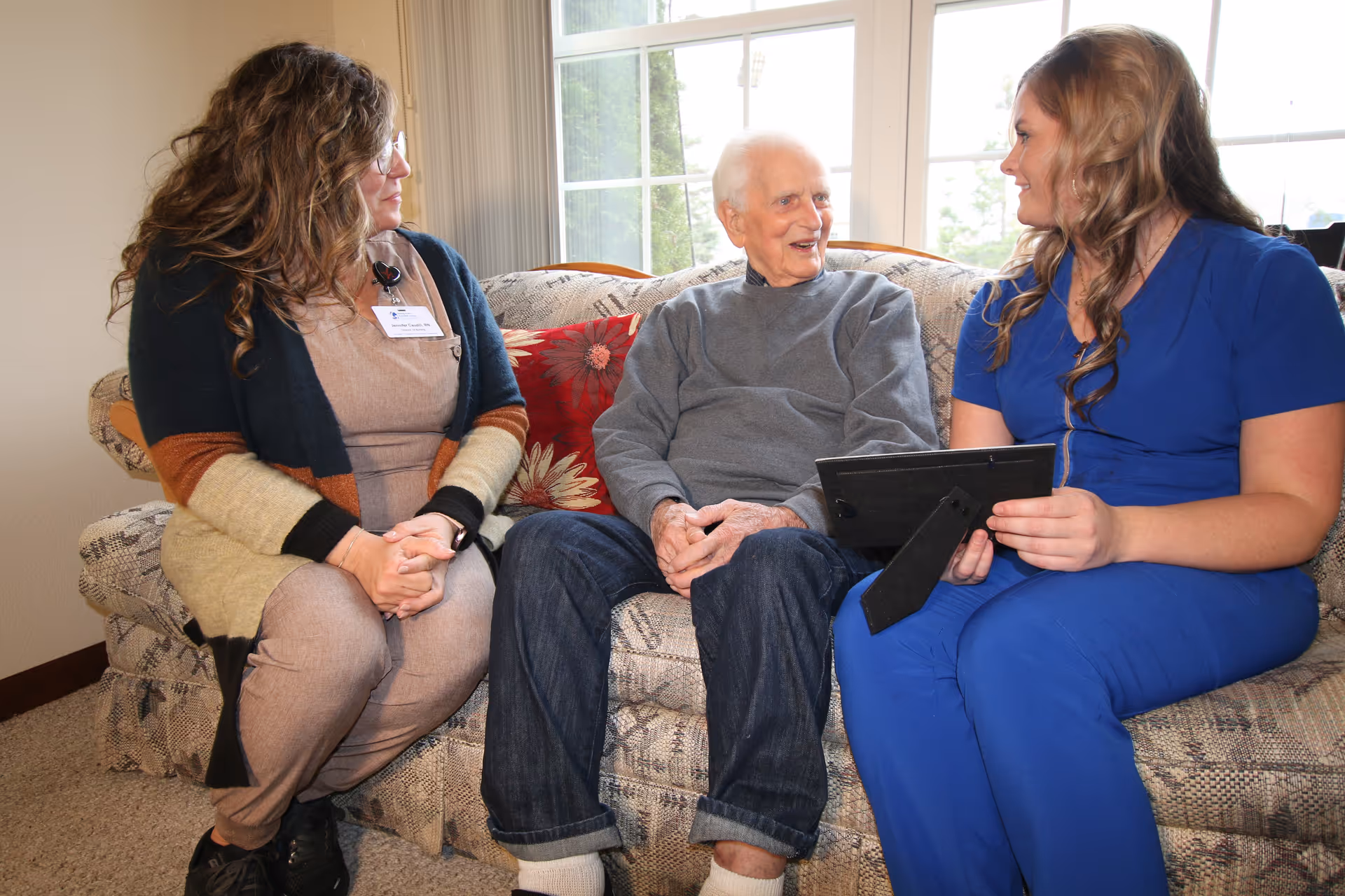 Three people sit on a patterned sofa in a bright living room.