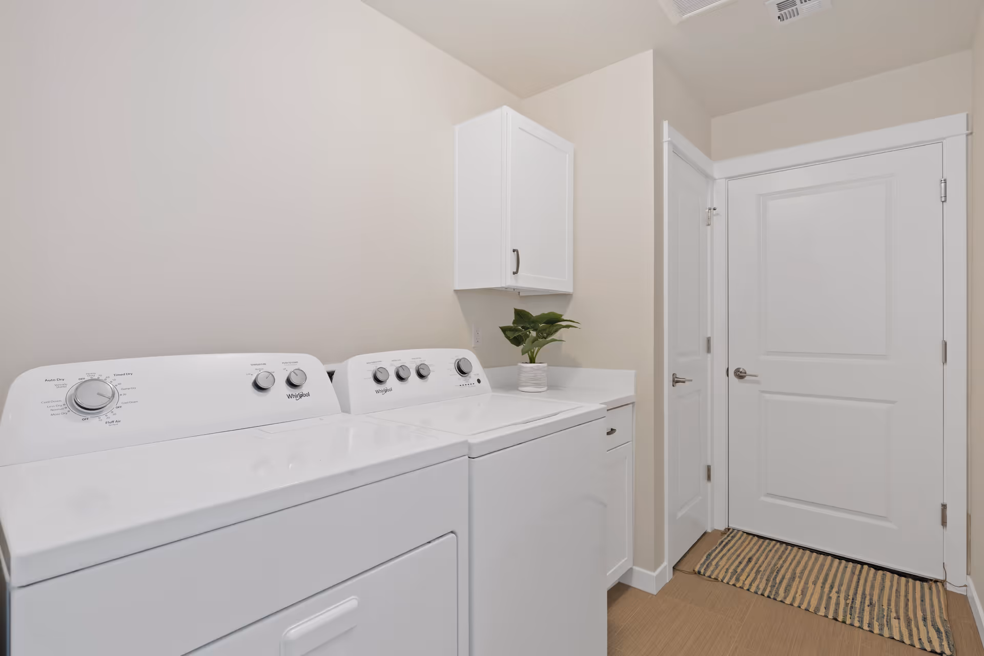 Laundry room with a white washing machine and dryer side by side, a white cabinet mounted on the wall above a countertop with a small green potted plant, and two white doors with silver handles. A striped rug is placed on the floor in front of the doors.