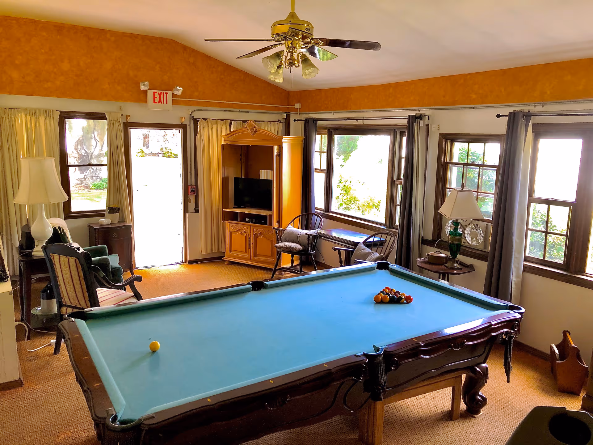 A cozy room with a pool table in the center, a ceiling fan with lights above, and several windows letting in natural light. The room has a TV cabinet, a couple of chairs, a small table, and lamps. The walls are painted in warm tones and there is an exit door visible.