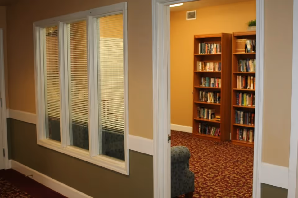 Interior view of a hallway with beige and olive green walls, white trim, and a carpet with a red and beige pattern. There is a window with blinds on the left wall and an open doorway on the right leading to a room with bookshelves filled with books and a blue patterned armchair.