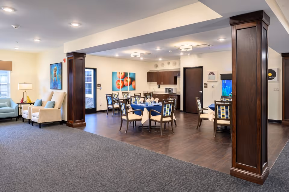 Open interior common area with dining tables set for meals, a seating lounge with armchairs, and dark wooden columns.