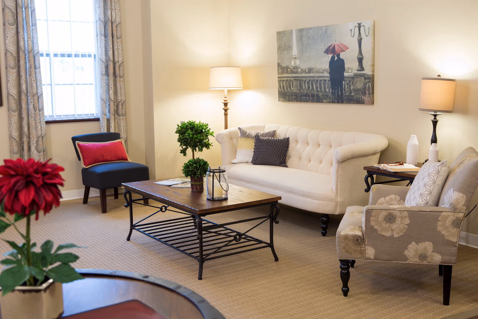 A bright living room with a tufted cream sofa, patterned armchair, coffee table, lamps, and wall art.