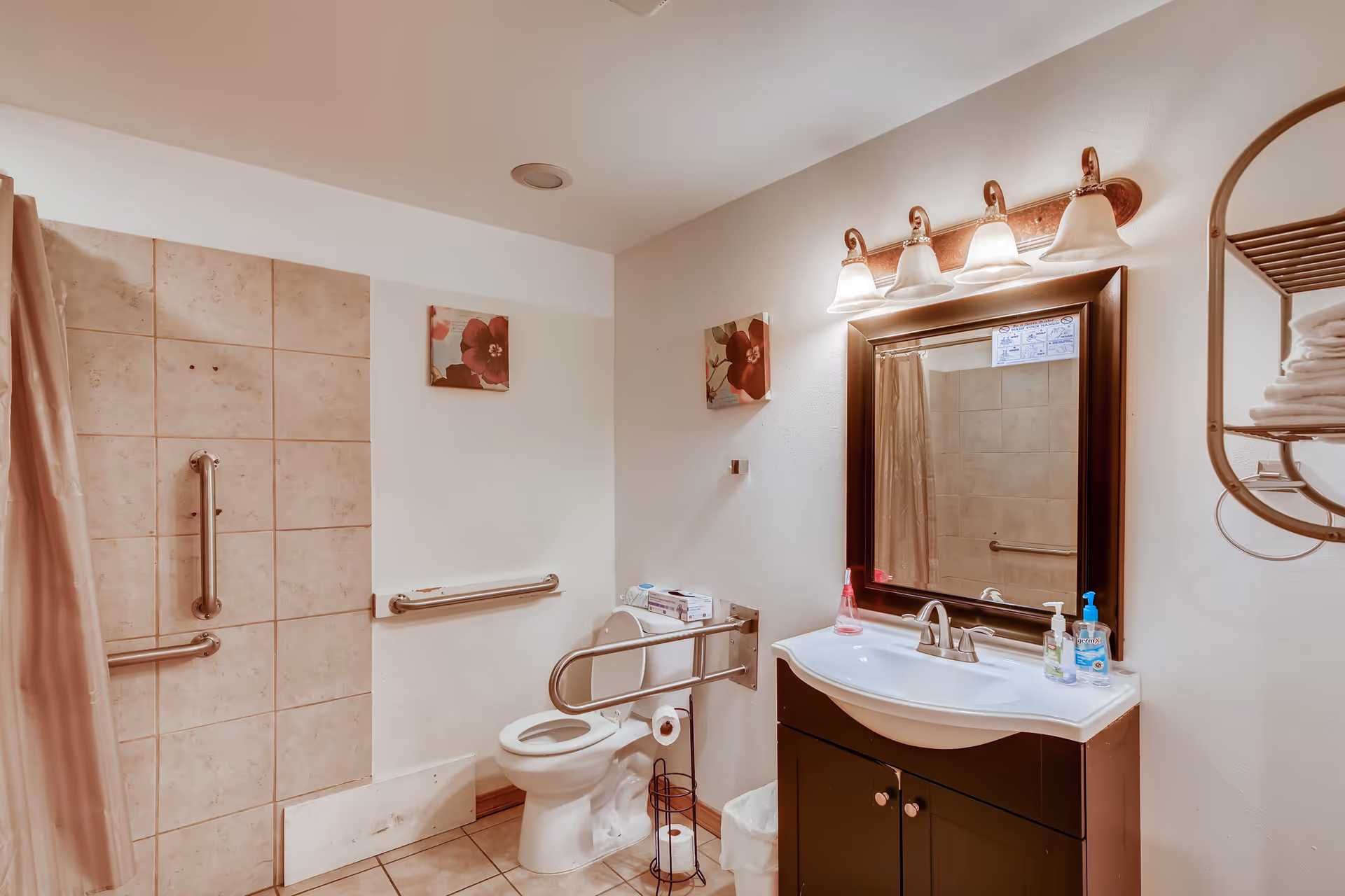 A clean bathroom with beige tiled shower area equipped with grab bars, a white toilet with a safety rail, a dark wood vanity with a white sink, a large mirror above the sink with four light fixtures, and a metal rack holding folded white towels on the wall.